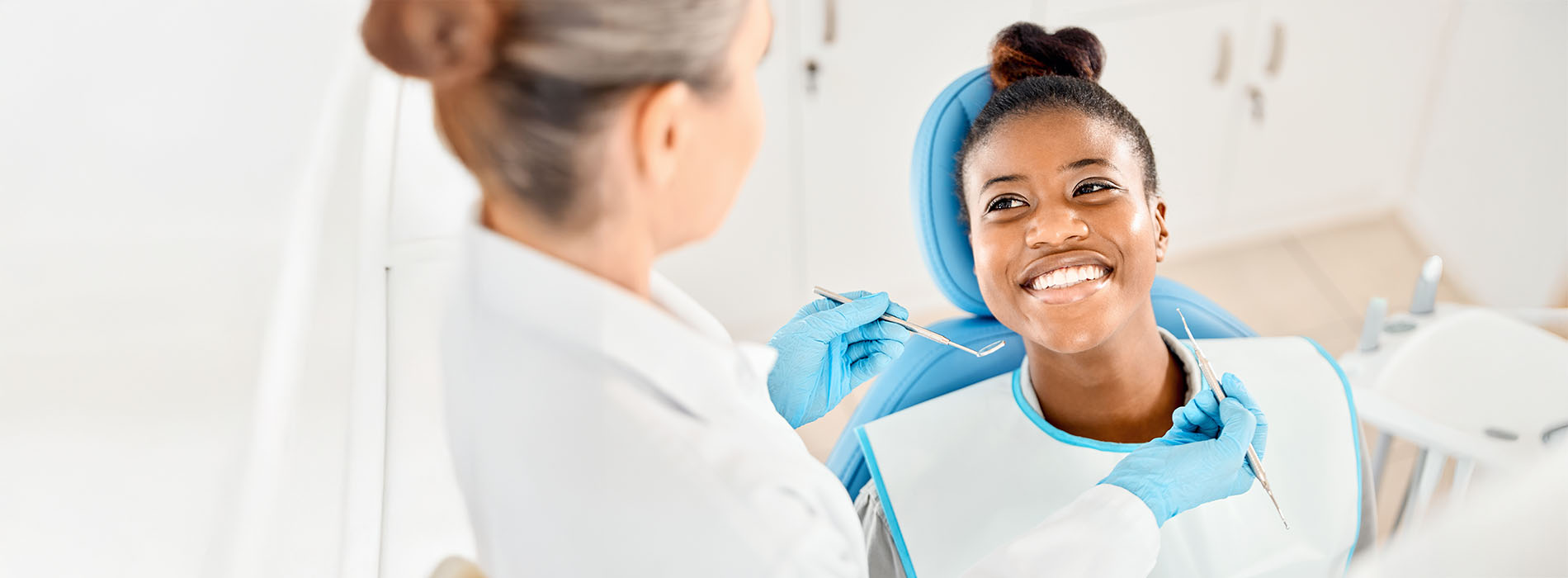 The image shows two individuals in a dental office setting, with one person seated in a dental chair receiving care from another standing behind, likely a dentist or dental hygienist.