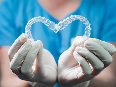 An image featuring a person s hands holding a pair of clear dental implants with a heart shape at their center, against a background that appears to be a healthcare setting with medical equipment and supplies.