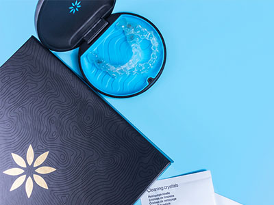 The image shows a closed box with a toothbrush inside, placed next to an open blue container, possibly containing dental floss, on a light blue background.