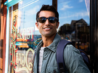 A man wearing sunglasses and a backpack stands confidently outside a storefront with a vintage sign, smiling towards the camera.