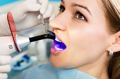 A woman receiving dental care with a blue light device, attended by a dentist using a mirror and a tool.