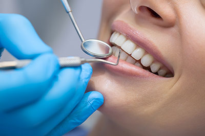 The image shows a dental professional performing a dental procedure on a patient s teeth, with the patient wearing a blue surgical gown and a smiling expression, while a dental hygienist uses dental tools and holds a tray of instruments.