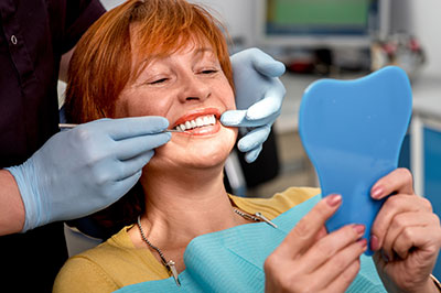 A woman is sitting in a dental chair with her mouth open, holding a blue dental model, while a dentist works on her teeth  they are both wearing face masks and gloves.