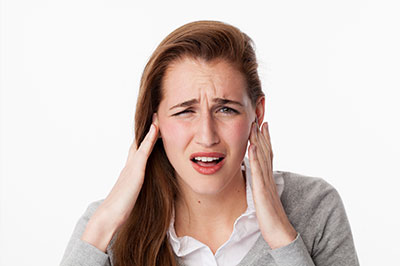 A woman with blonde hair, wearing a grey sweater, holds her hand on her head, looking distressed, with an  alt  tag description of  a woman experiencing discomfort or pain.