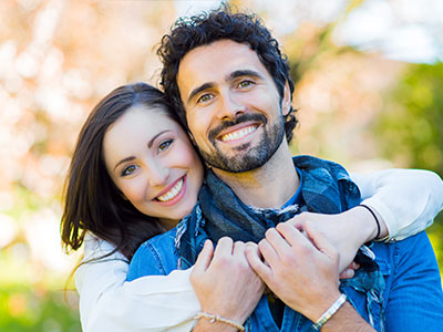 The image depicts a man and a woman embracing each other outdoors  the man has facial hair and is wearing a blue top, while the woman is smiling and has her arms around him. They appear to be in a joyful moment together.