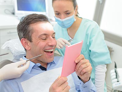 The image shows a man sitting in a dental chair with a smiling expression, holding up a pink card with a surprised look on his face. A woman wearing a surgical mask is standing behind him, looking at the card with a concerned expression.