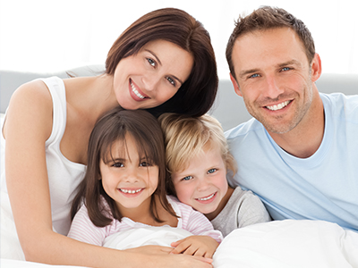 The image shows a family of four, including two adults and two children, all smiling and posing together on a bed with white sheets.