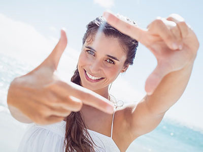 A woman with long hair, wearing a white top, smiles at the camera while holding up two fingers.